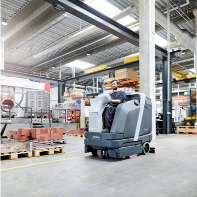 A Nilfisk SC6000 Ride-on Floor Scrubber Dryer being operated in a warehouse.